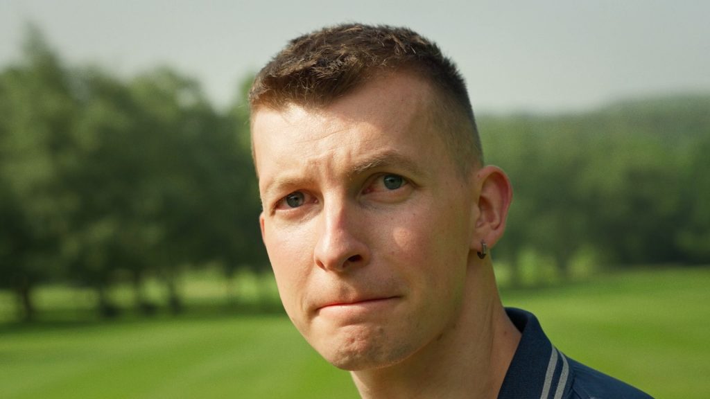 A still from the film Grasshoper Par3. A man on a golf course looks tentatively towards the camera.